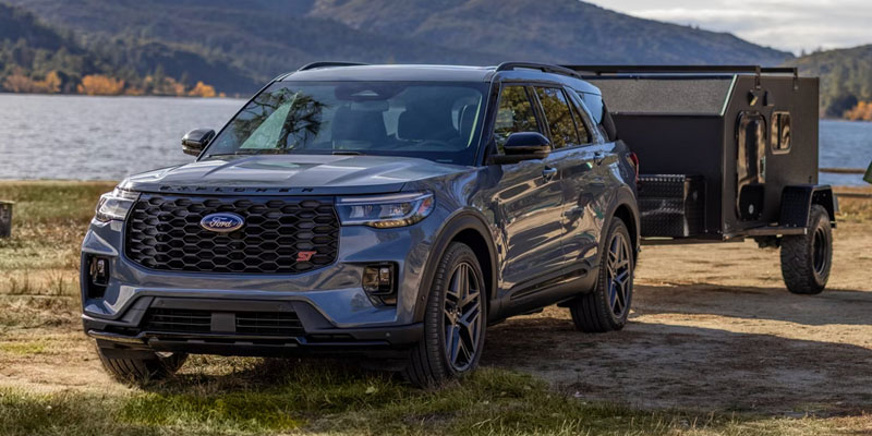Blue Ford Explorer SUV parked near a lake, towing a black trailer, with trees and hills in the background