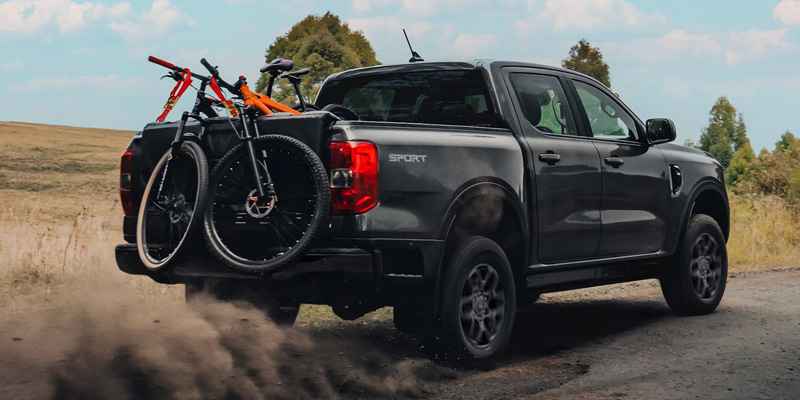 Gray Ford Ranger driving on a dirt road with two bicycles mounted on the tailgate, kicking up dust in a rural landscape
