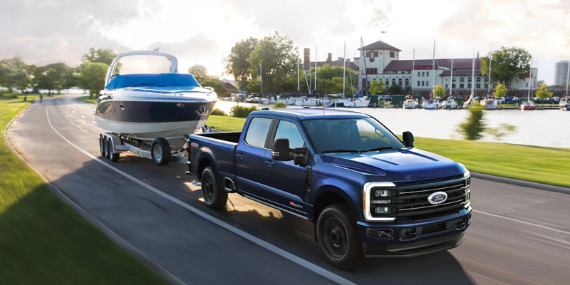 Blue Ford Super Duty pickup truck towing a boat on a trailer along a paved road beside a waterfront marina, with buildings and boats visible in the background.
