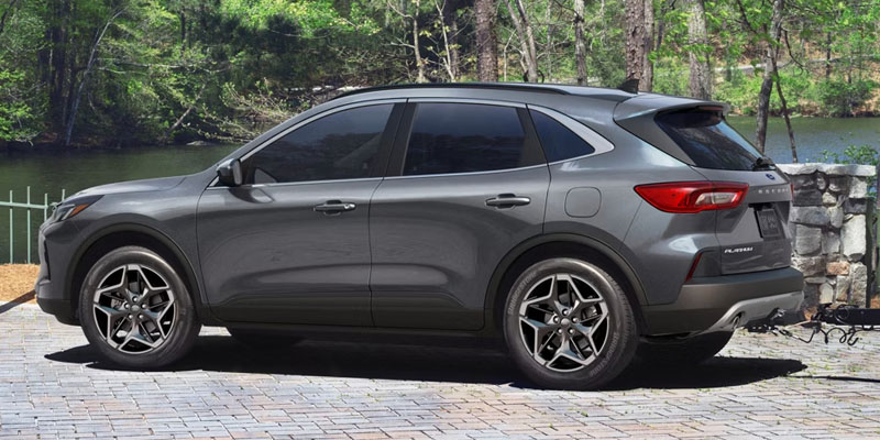 Side profile view of a gray Ford Escape parked on a paved driveway near a wooded lakeside setting, showcasing its exterior design, wheels, and rear lighting.