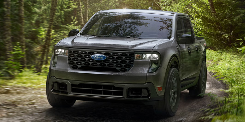 Front view of a gray Ford Maverick truck driving on a narrow forest trail, surrounded by dense green trees and foliage, highlighting off-road capability.