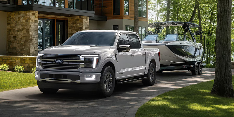 White Ford F-150 truck towing a large boat on a trailer along a paved driveway in a wooded lakeside residential setting near a modern home.